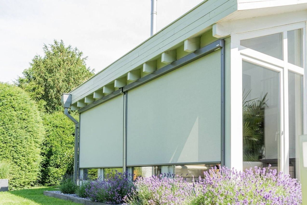 Hausfassade mit zwei heruntergelassenen Außenrollos an großen Fenstern und Garten mit Sträuchern und Blumen