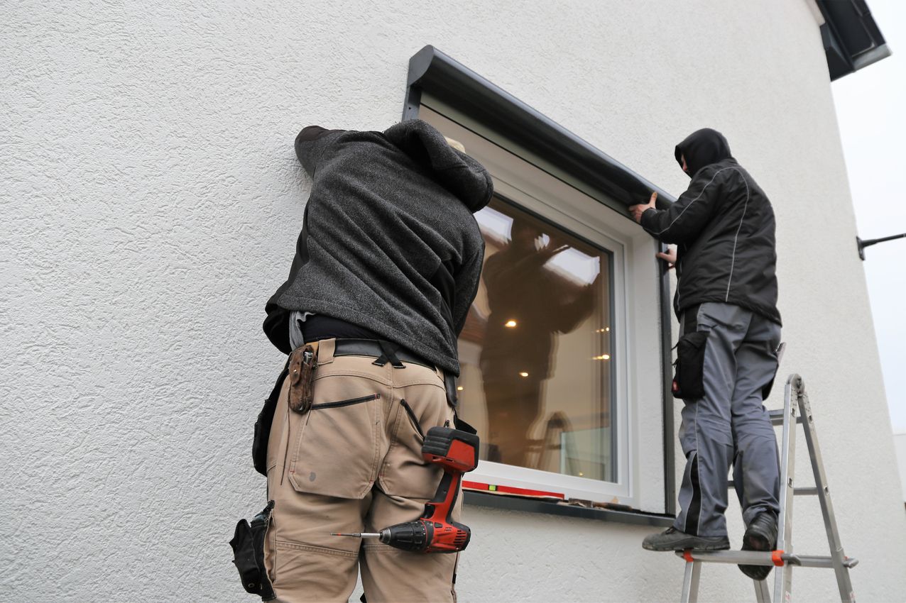 Zwei Handwerker montieren ein Fenster an einer weißen Hauswand, einer steht auf einer Leiter, der andere auf dem Boden