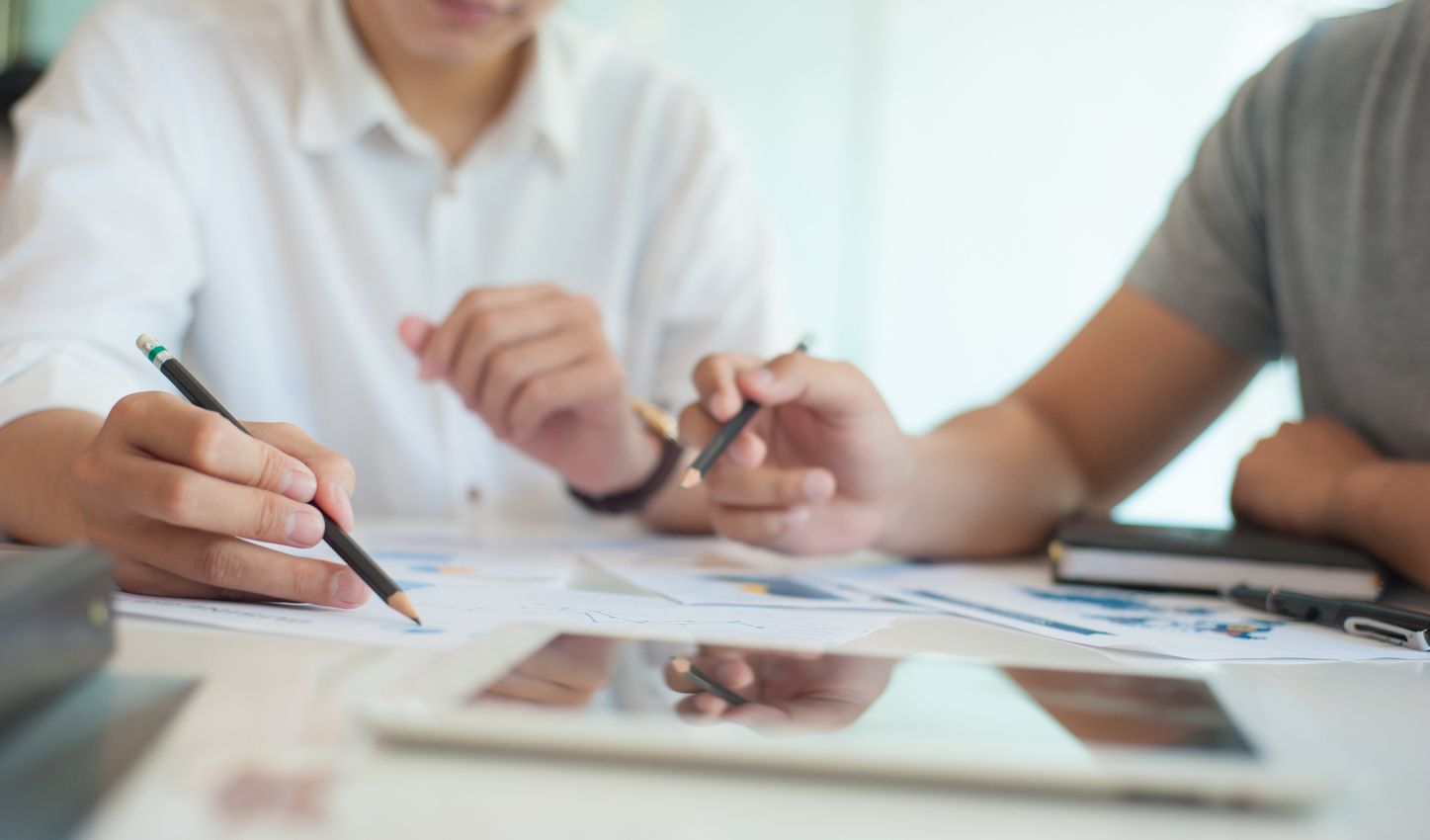 Nahaufnahme von zwei Personen, die an einem Tisch sitzen und mit Stiften auf Papier zeichnen, im Vordergrund liegt ein Tablet